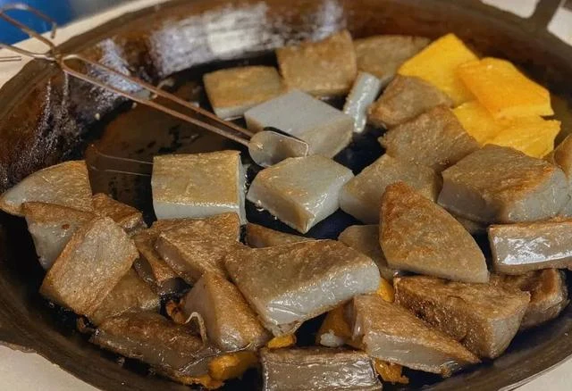Grey-brown Lijiang jidou jelly pieces in a black cooking pan with a spatula.