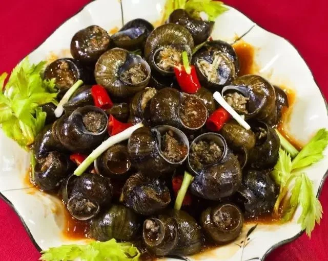 Guilin-style stuffed river snails with visible meat filling in dark shells, red chili and spring onion on a white plate; published by Guangxi Daily in 2018. The source does not identify the restaurant or original photographer.