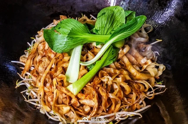 Geji braised pancake strips with meat, bean sprouts and green bok choy in a wok; Zhengzaiban photograph published by Zhengzhou Daily in 2023, with the original watermark retained.